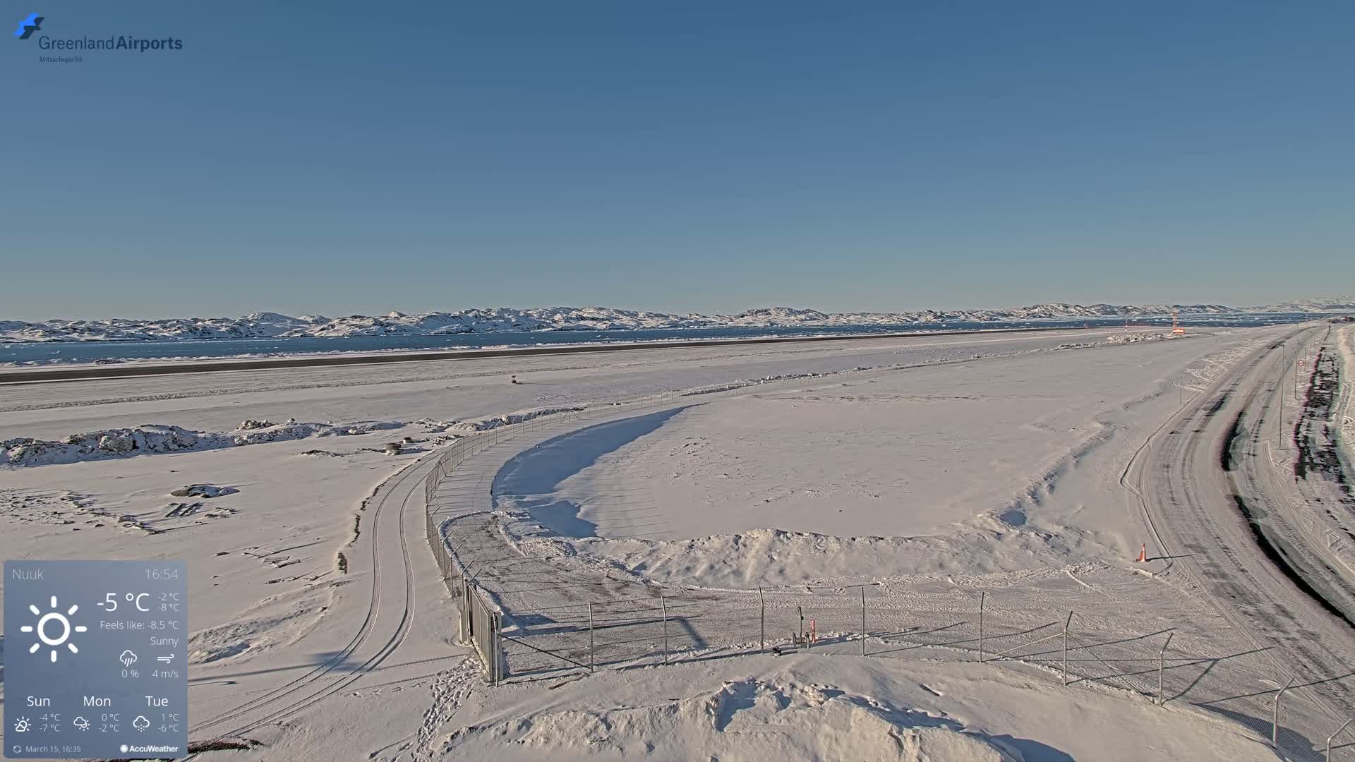 A snow-covered airport runway and surrounding area extend to a body of water and distant, snow-capped mountains under sunny skies.