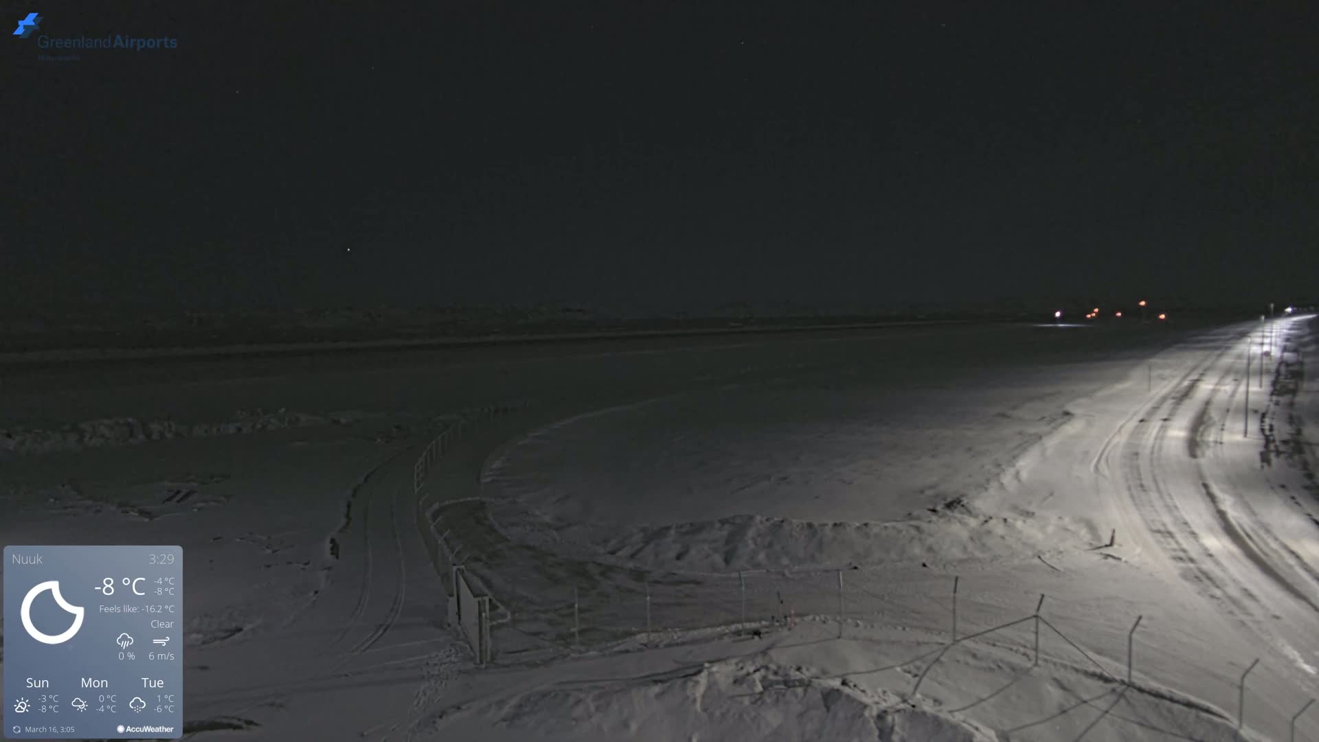 A snow-covered, nighttime scene shows a road curving through a flat, snow-covered expanse under a clear sky.