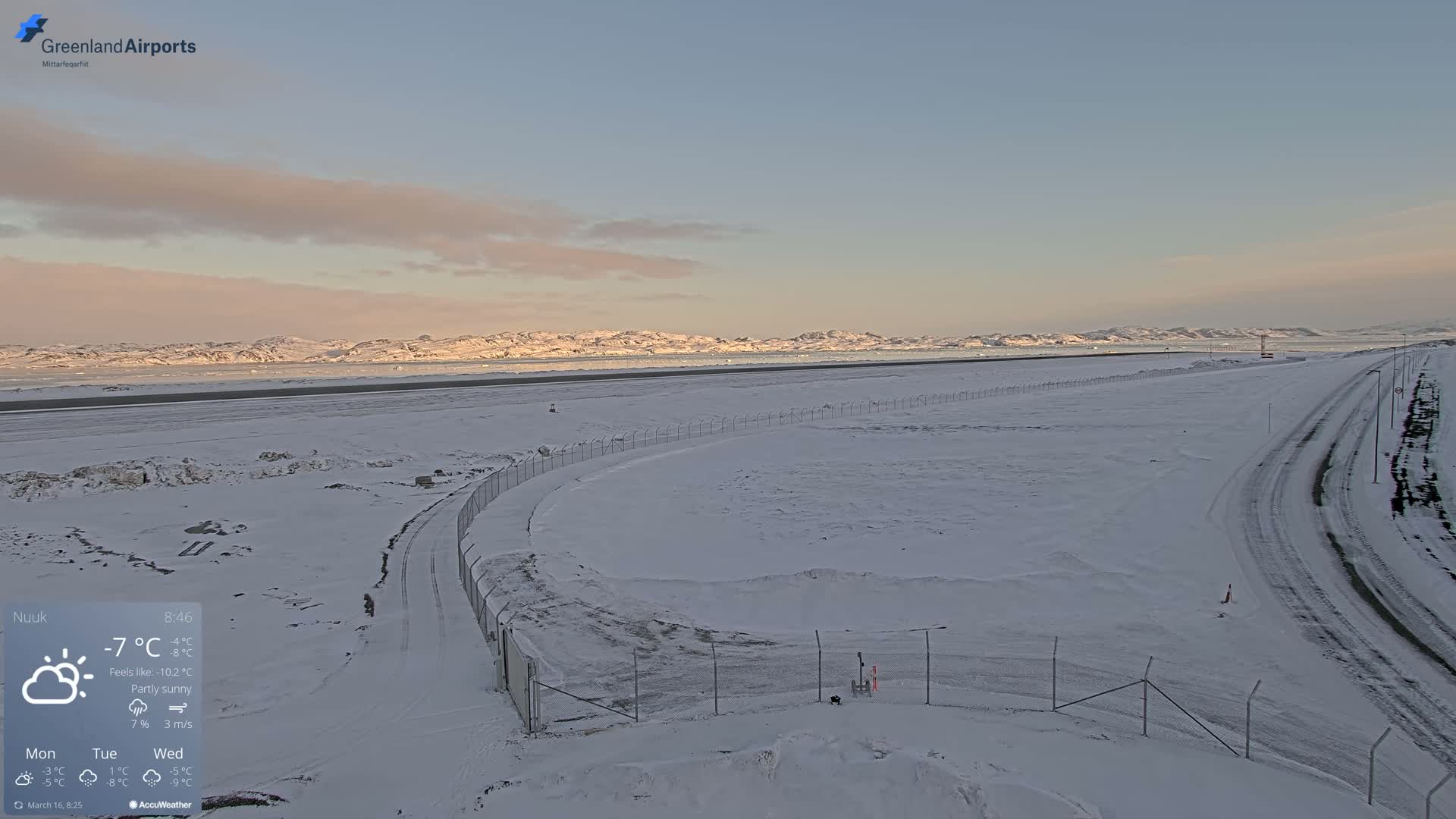 A snow-covered airfield with a chain-link fence and roads stretches toward a partly sunny, distant mountain range under a pale sky.