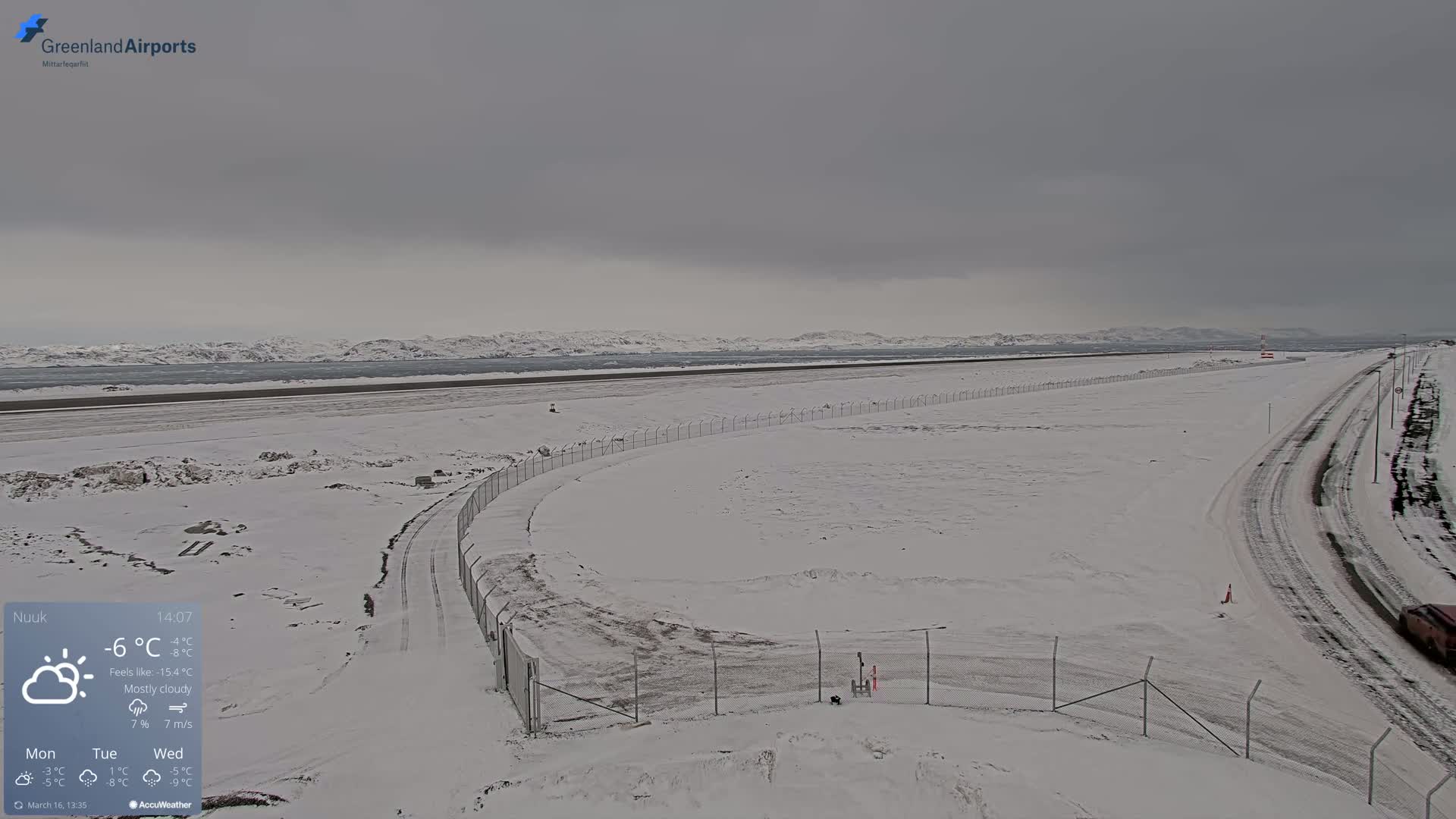 A snow-covered airport runway and surrounding area extend to a mostly cloudy, distant, snow-covered mountain range.