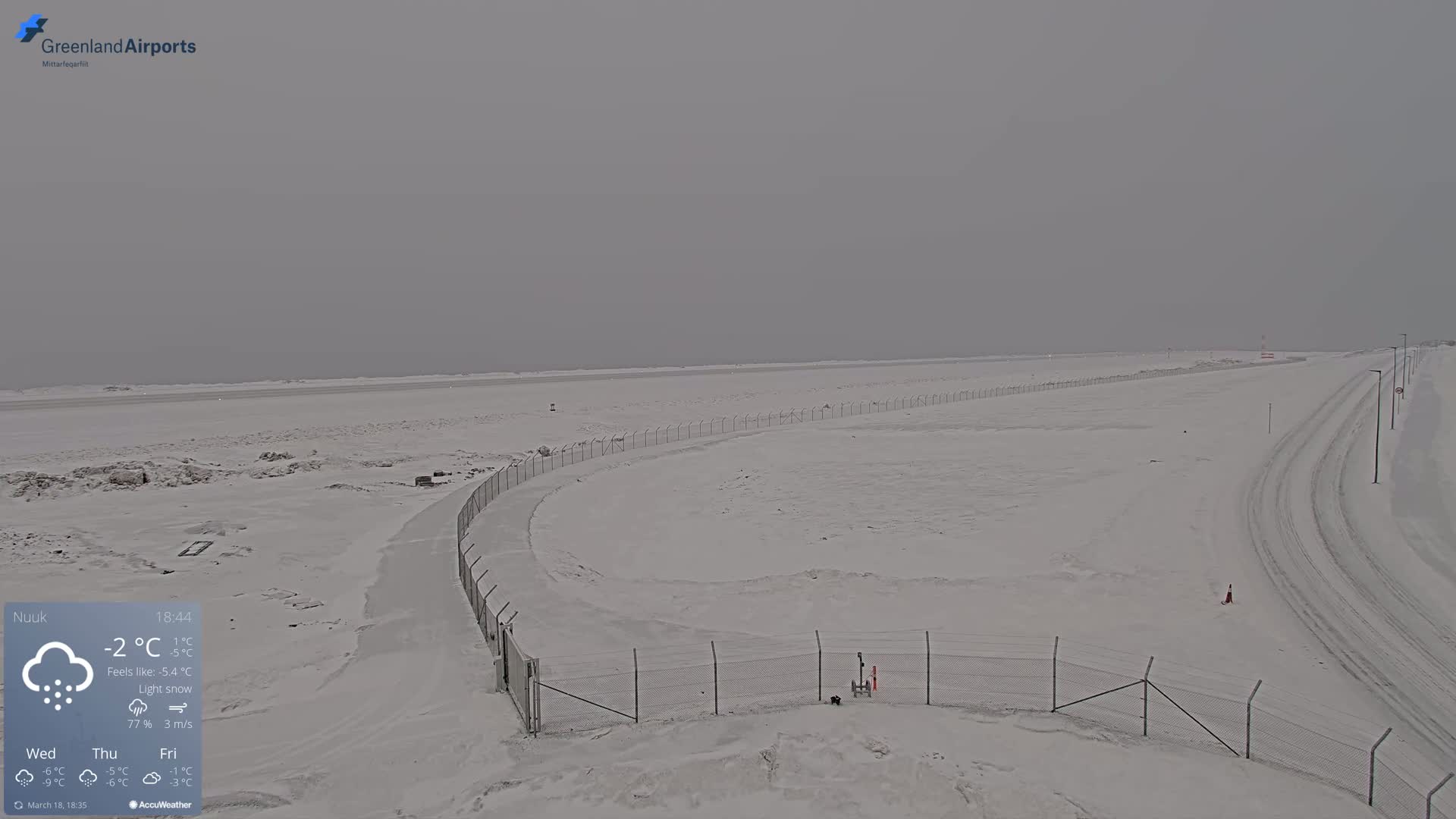 A snow-covered airfield with a chain-link fence and light snow falling under an overcast sky.