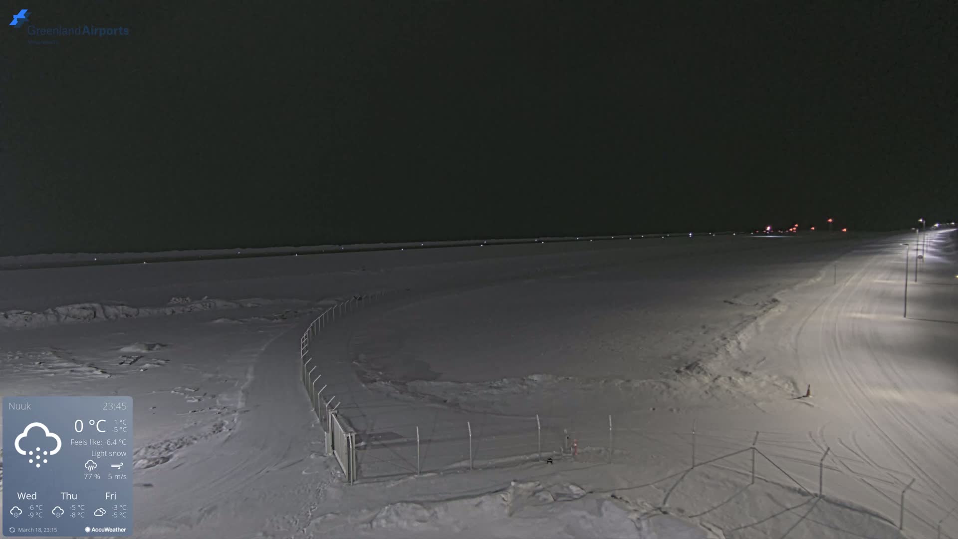 A snow-covered airfield is illuminated at night under conditions of light snow.