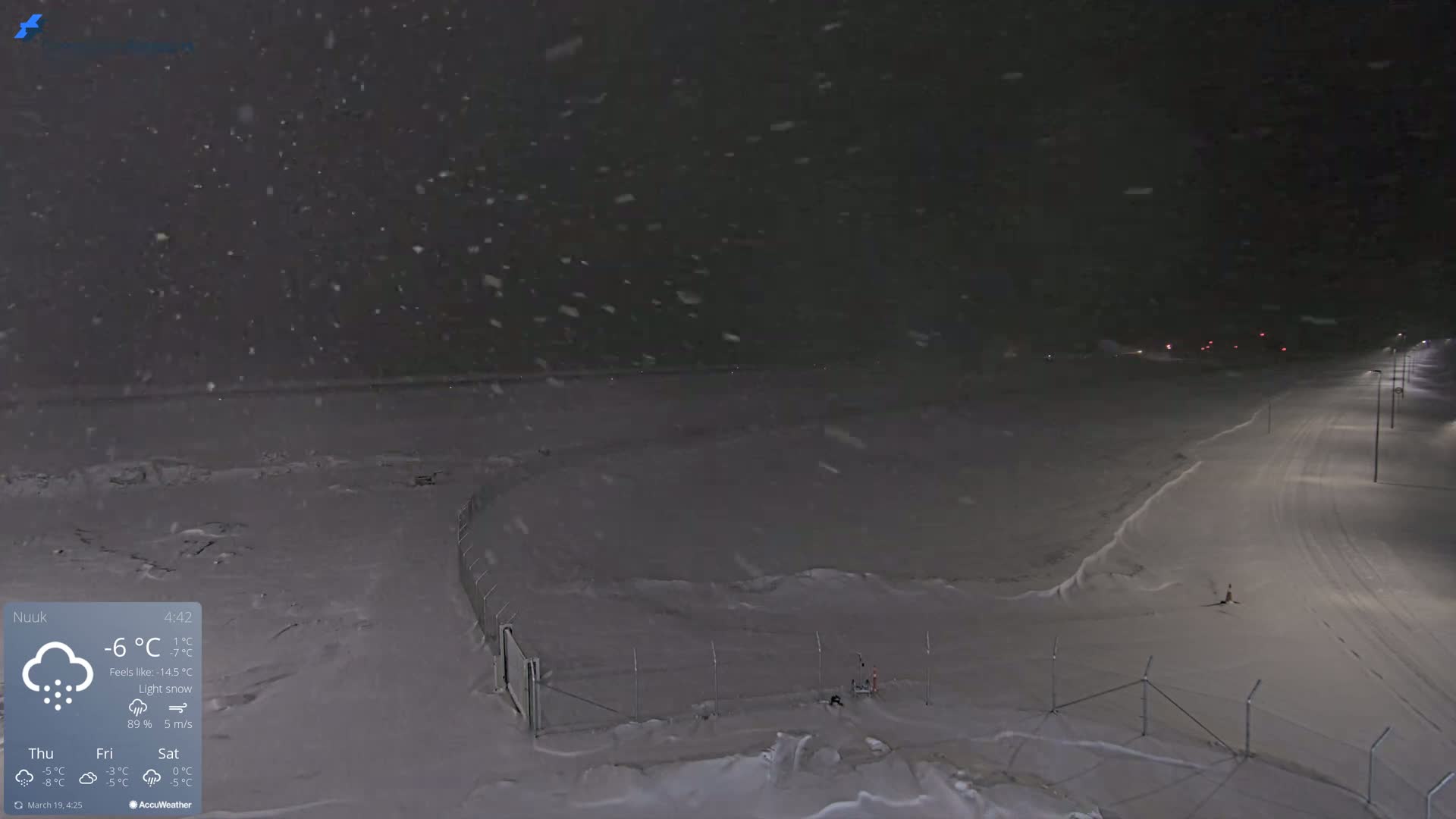 A snow-covered landscape at night, lightly snowing, with a road and fence visible under dim lighting.