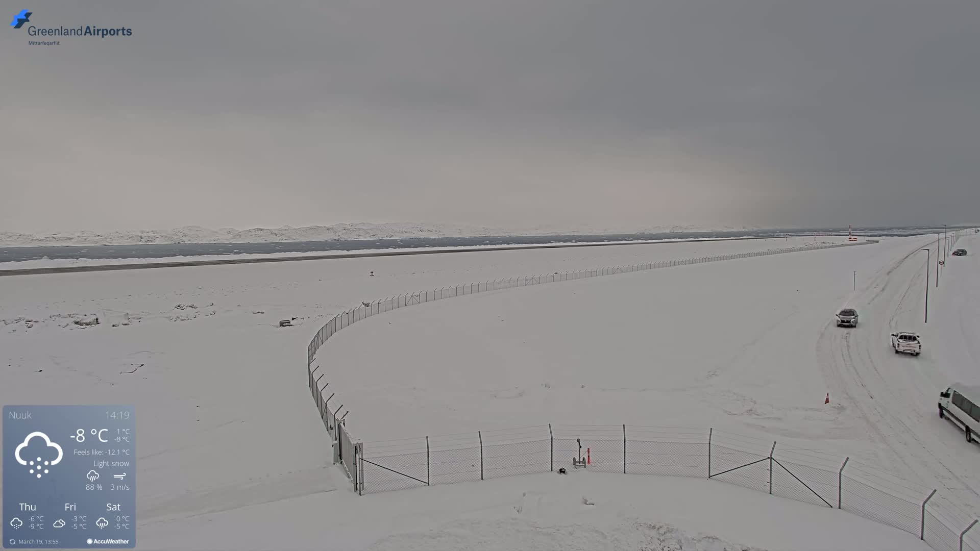 A snow-covered airfield with a chain-link fence and a few vehicles driving on a snow-covered road under a light snow.