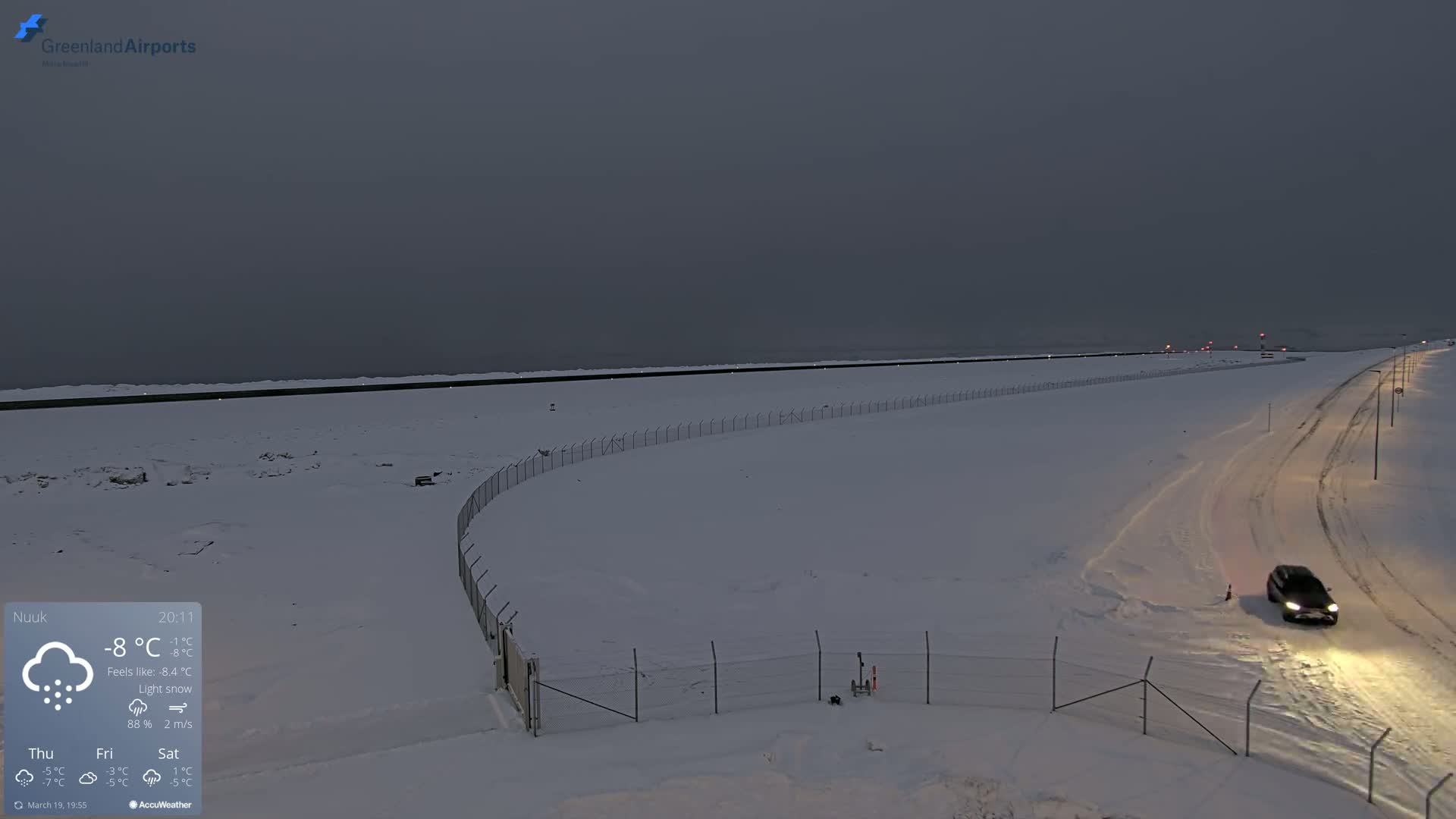 A snow-covered landscape with a road and fence under a dark, overcast sky, with light snow falling.