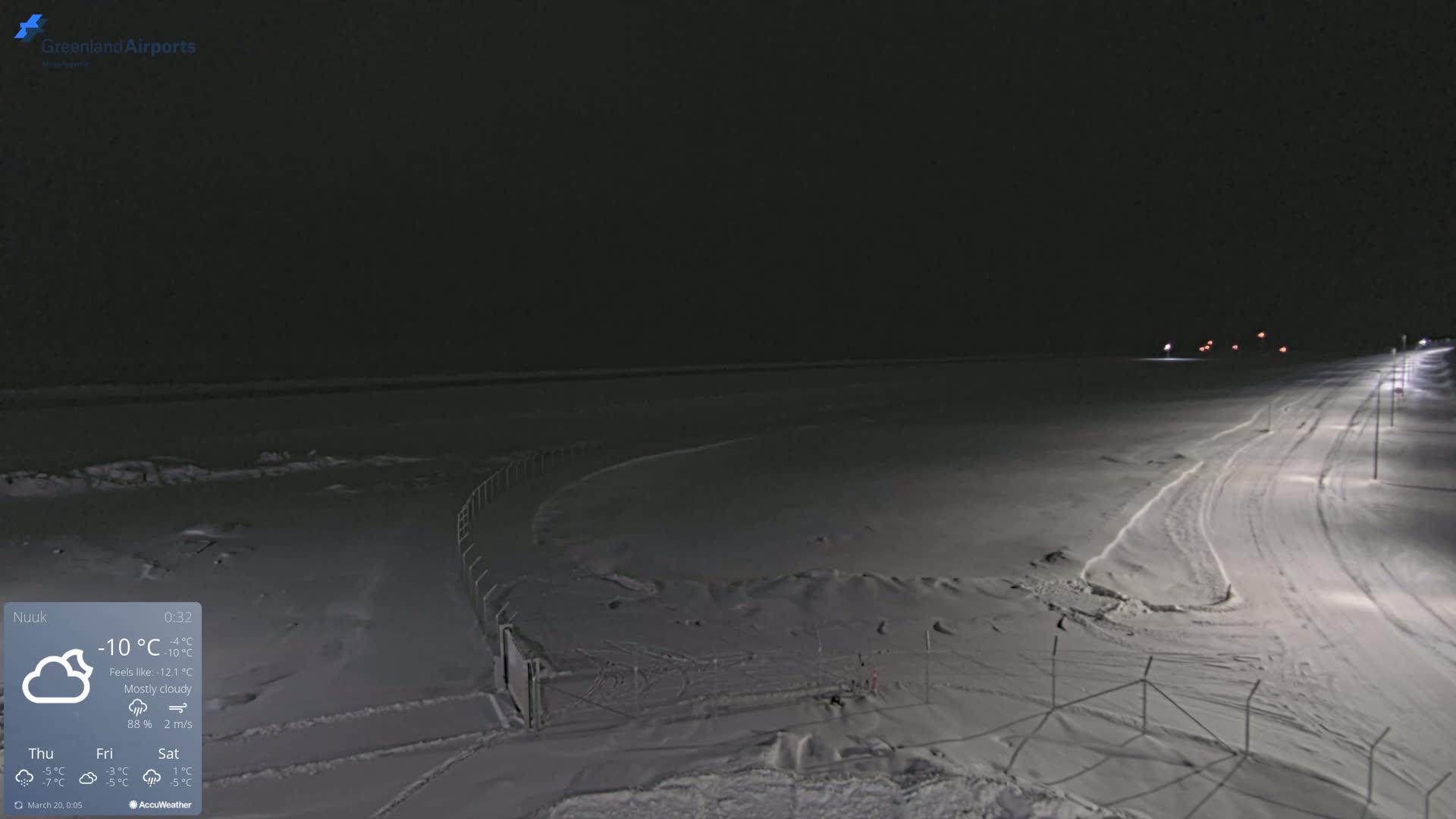 A snow-covered, dimly lit landscape at night, under mostly cloudy skies, with a road and fence visible.