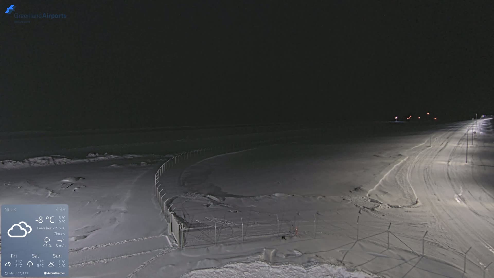 A snow-covered, dark nighttime scene shows a road and fence illuminated by lights, under cloudy conditions.