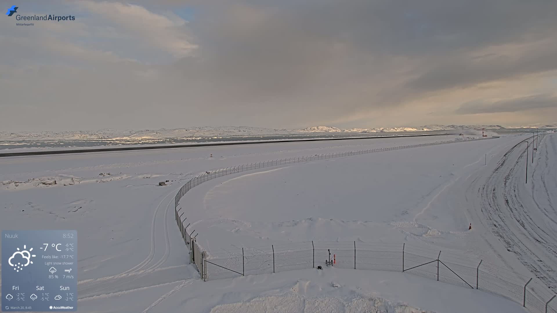 A snow-covered airport runway and surrounding area are visible under a light snow shower with overcast skies and distant, snow-capped mountains.