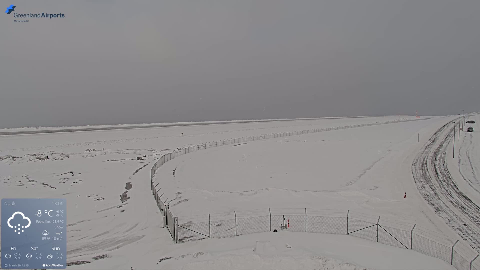 A snow-covered airport runway and access road are visible under a gray, overcast sky with light snowfall.