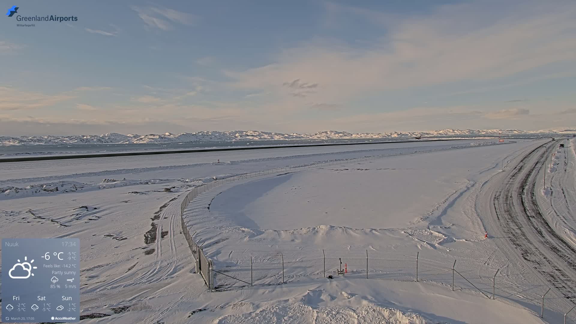 A snow-covered airport tarmac and roadway extend toward a partly sunny sky and distant snow-capped mountains.