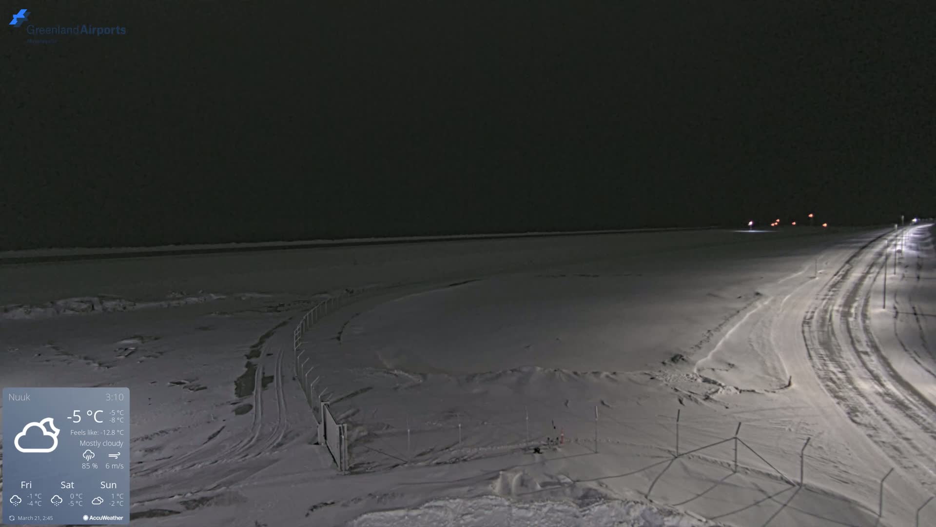 A snow-covered landscape at night under mostly cloudy skies, showing a road with lights and a fenced-in area.
