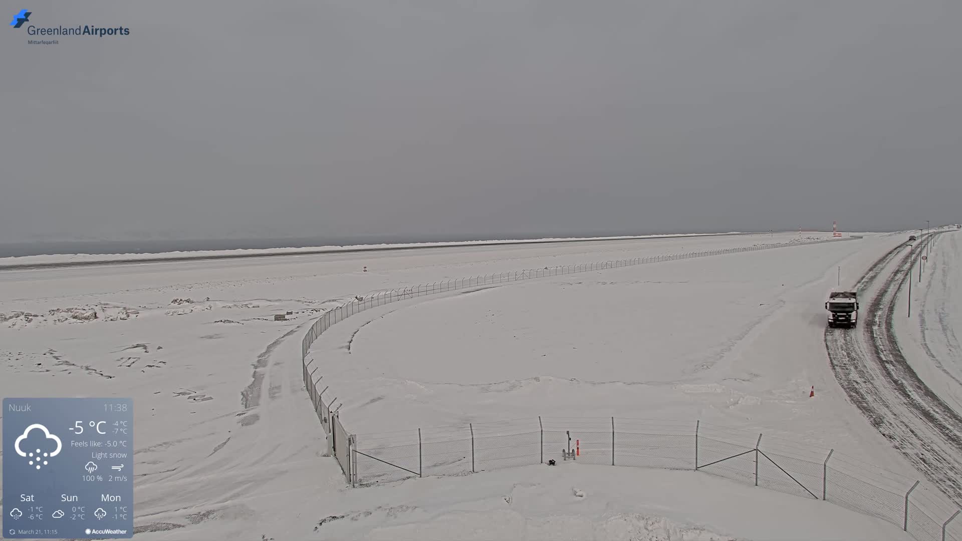 A snow-covered airport runway and surrounding area with a truck driving on a nearby road under light snow.