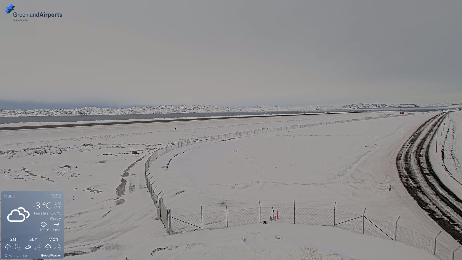 A snow-covered airport runway and access road extend toward a distant, snow-capped mountain range under a cloudy sky.