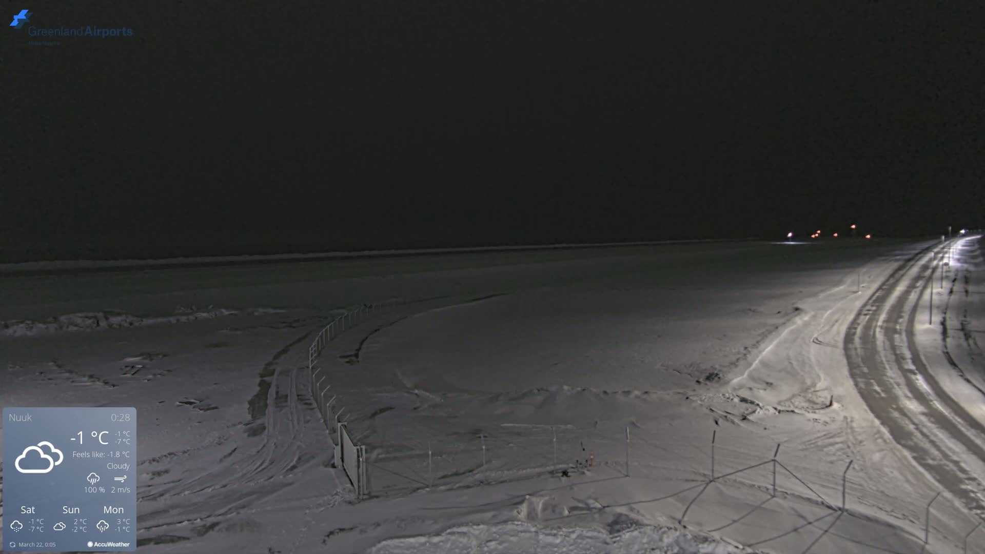 A snow-covered landscape at night under cloudy skies, with a road and fence visible.