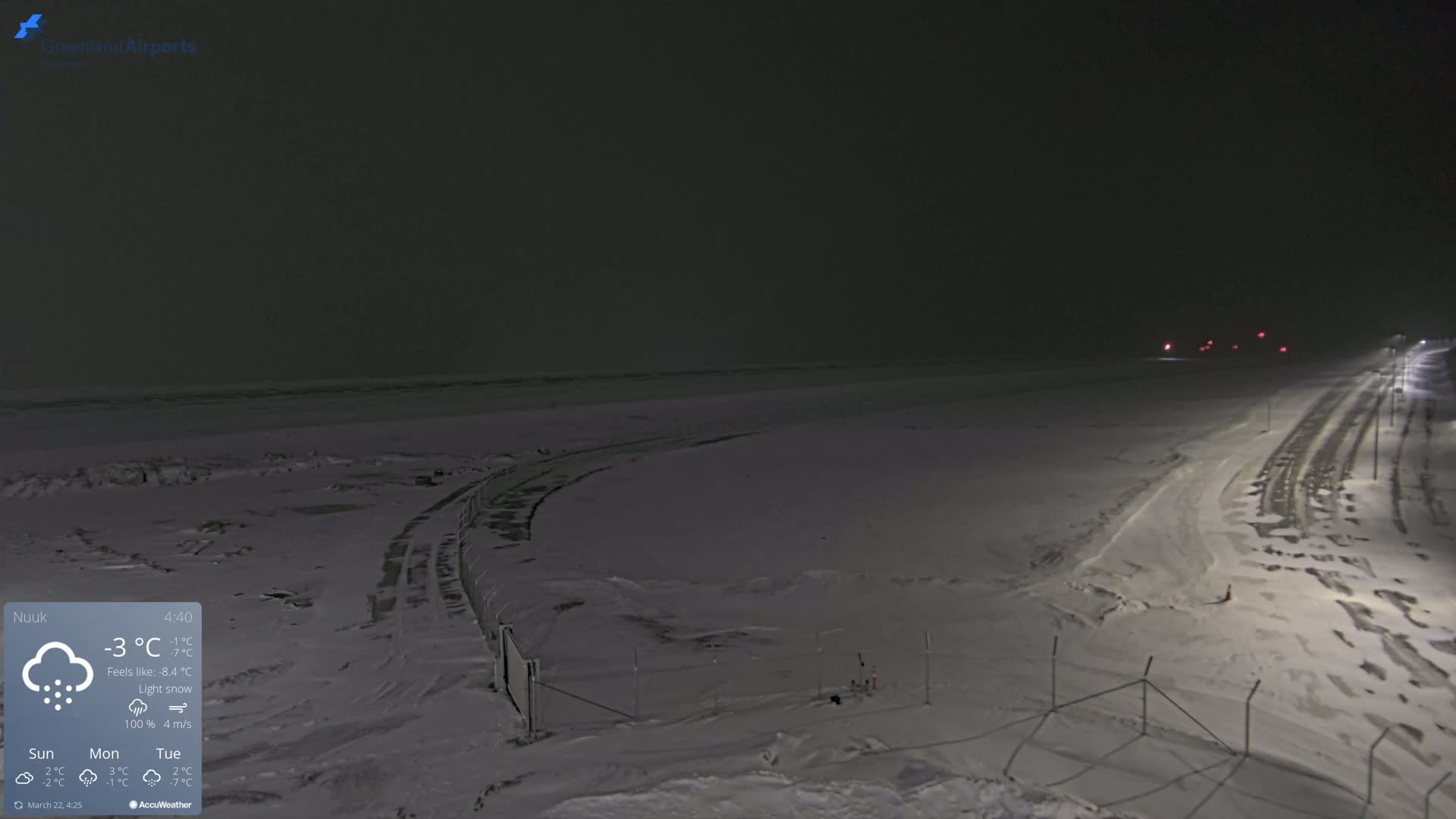 A snow-covered landscape at night, with a road and a fence visible under light snowfall.