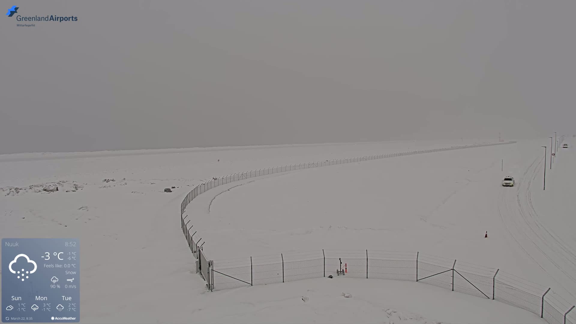 A snow-covered airfield is visible under a hazy sky, with a few vehicles traveling on a snow-covered road.