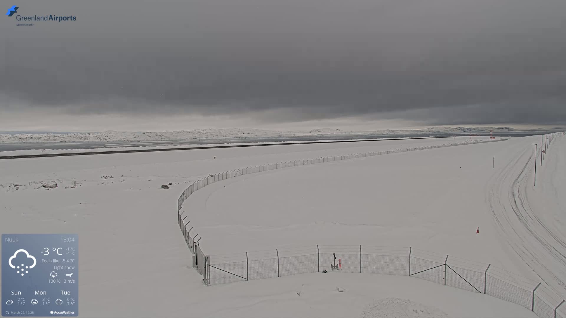 A snow-covered landscape with a chain-link fence curving through it, under a gray sky with light snow falling.
