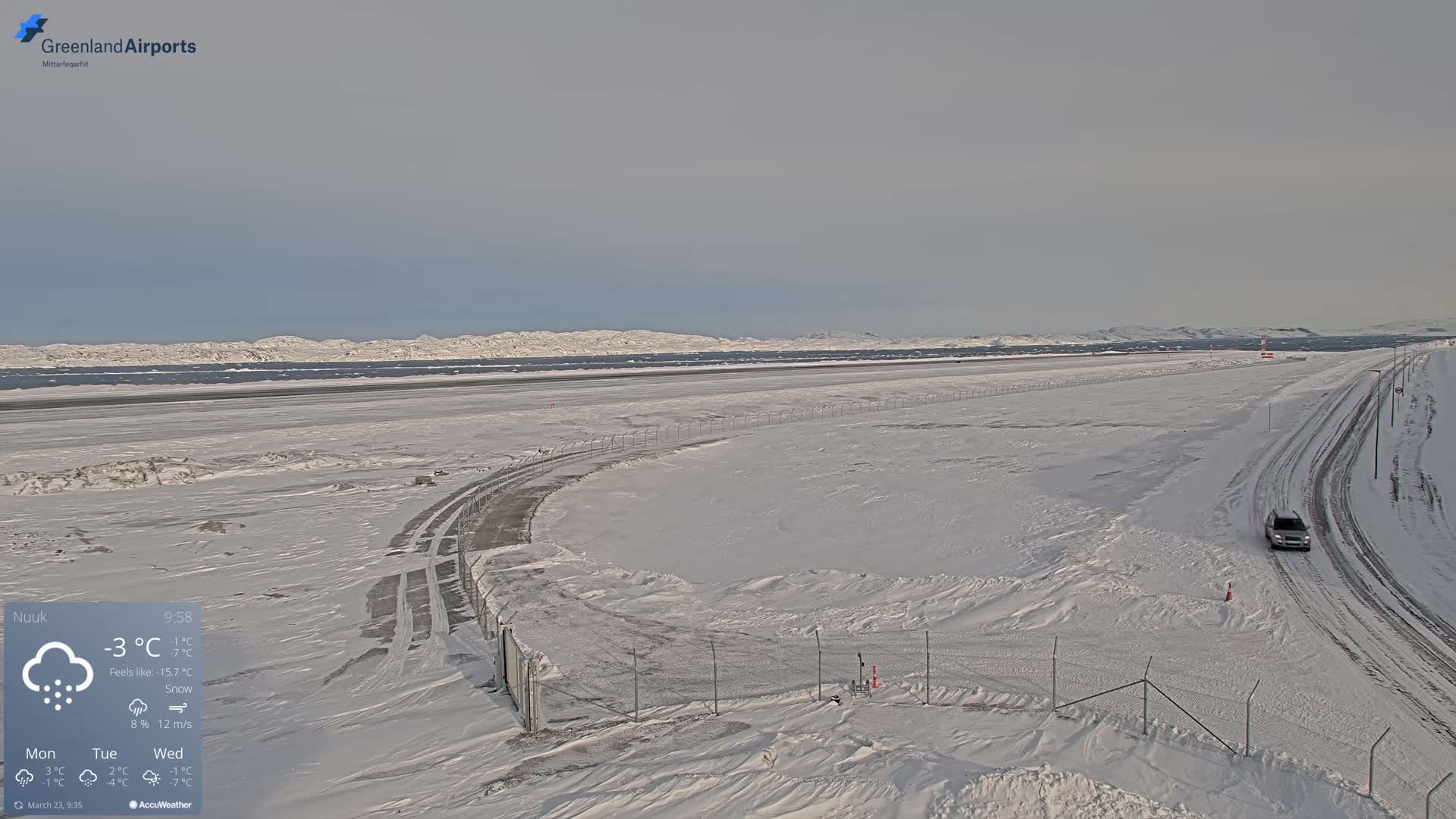 A snow-covered airport runway and access road extend to a distant coastline under a cloudy sky.