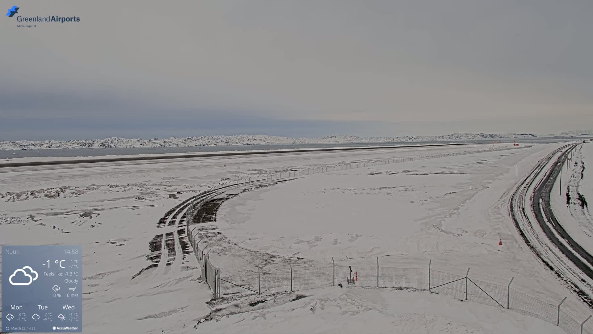 A snow-covered airport runway and surrounding area extend to a body of water backed by distant, snow-dusted mountains under a cloudy sky.