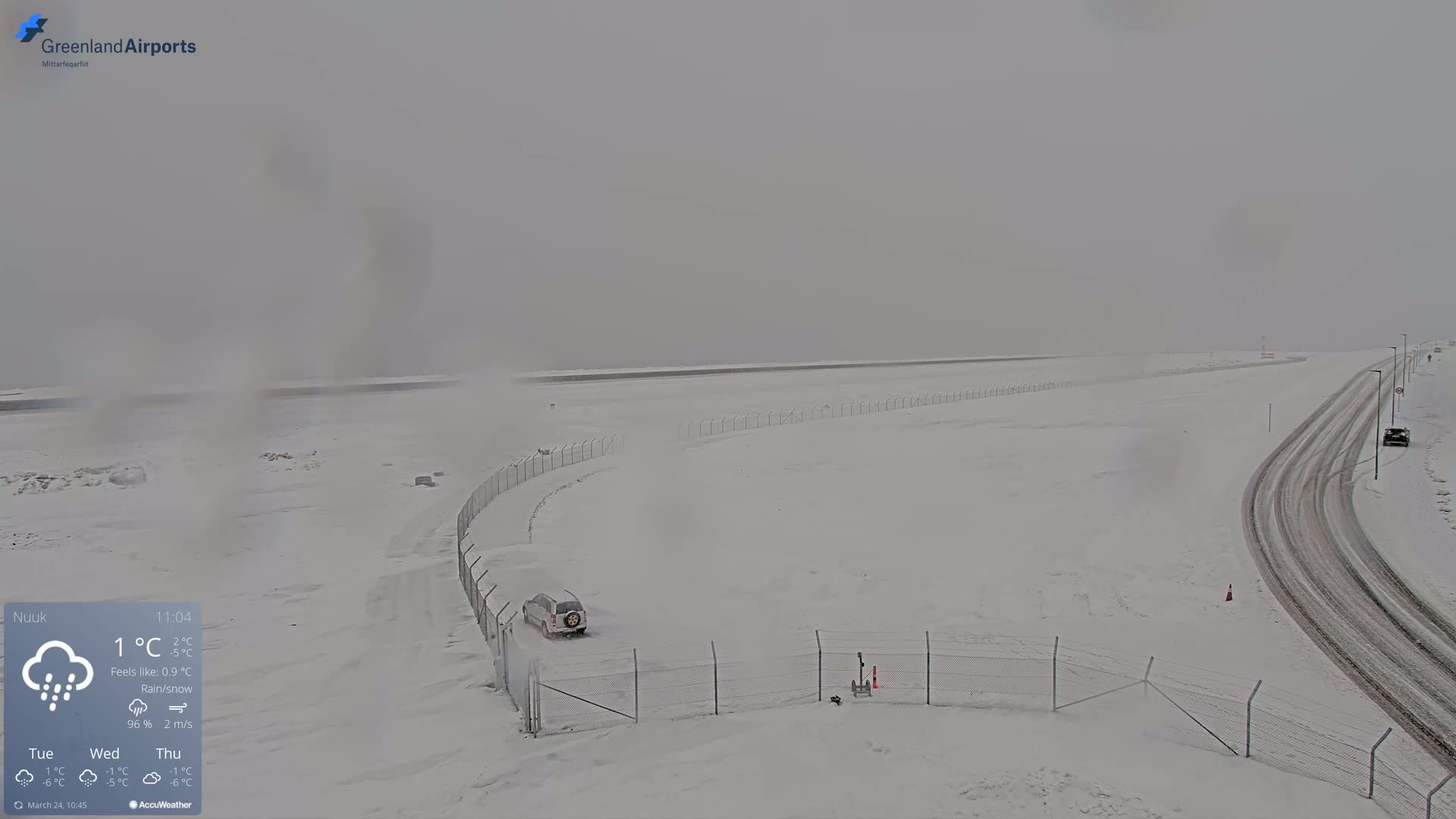 A snow-covered airport runway and adjacent roads are visible under overcast skies with rain and snow falling.