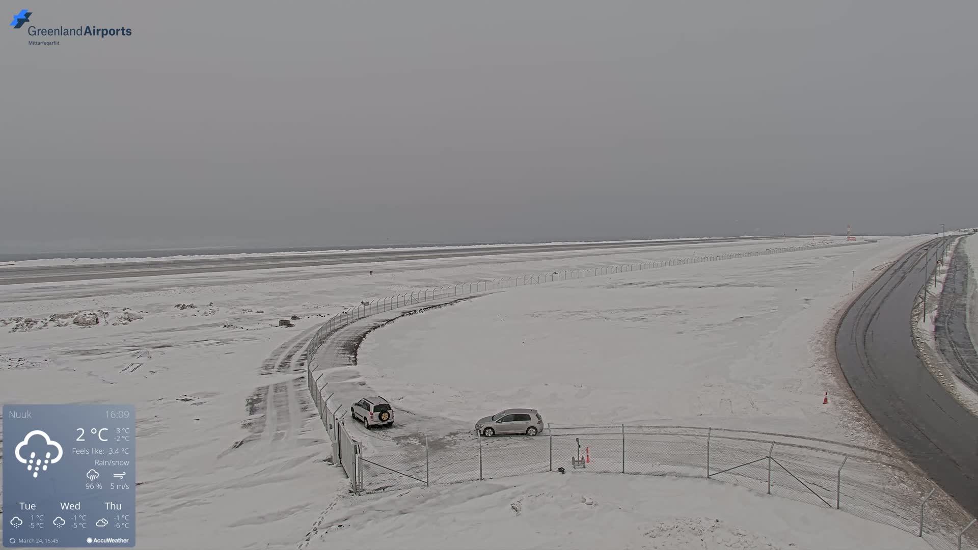 A snow-covered airport runway with two vehicles parked near a chain-link fence, under a gray, overcast sky with rain and snow.