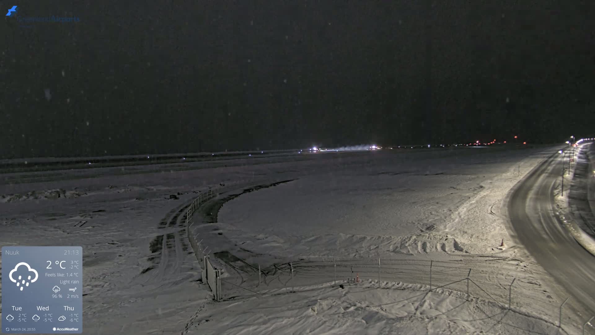 A snow-covered airport runway and road are visible at night under light rain.