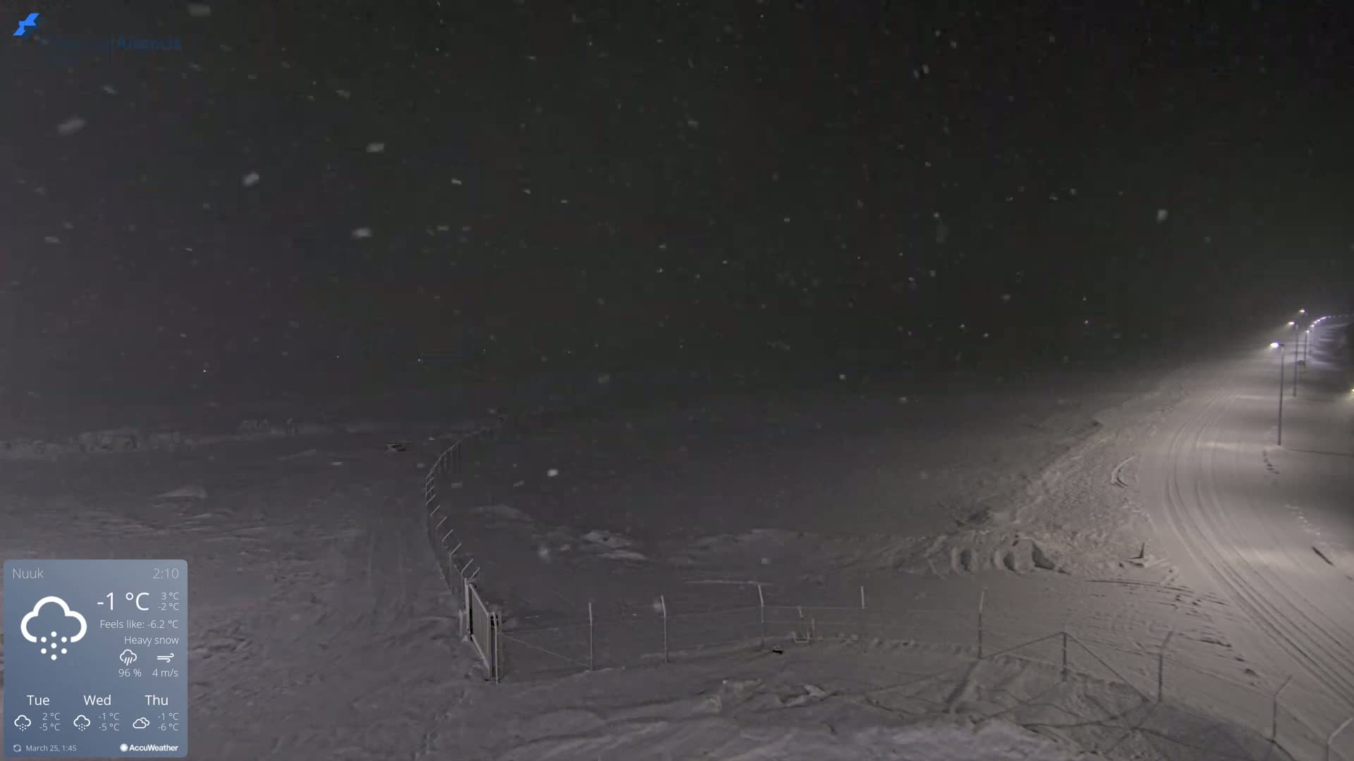 A snow-covered landscape at night is illuminated by streetlights, with heavy snow falling.