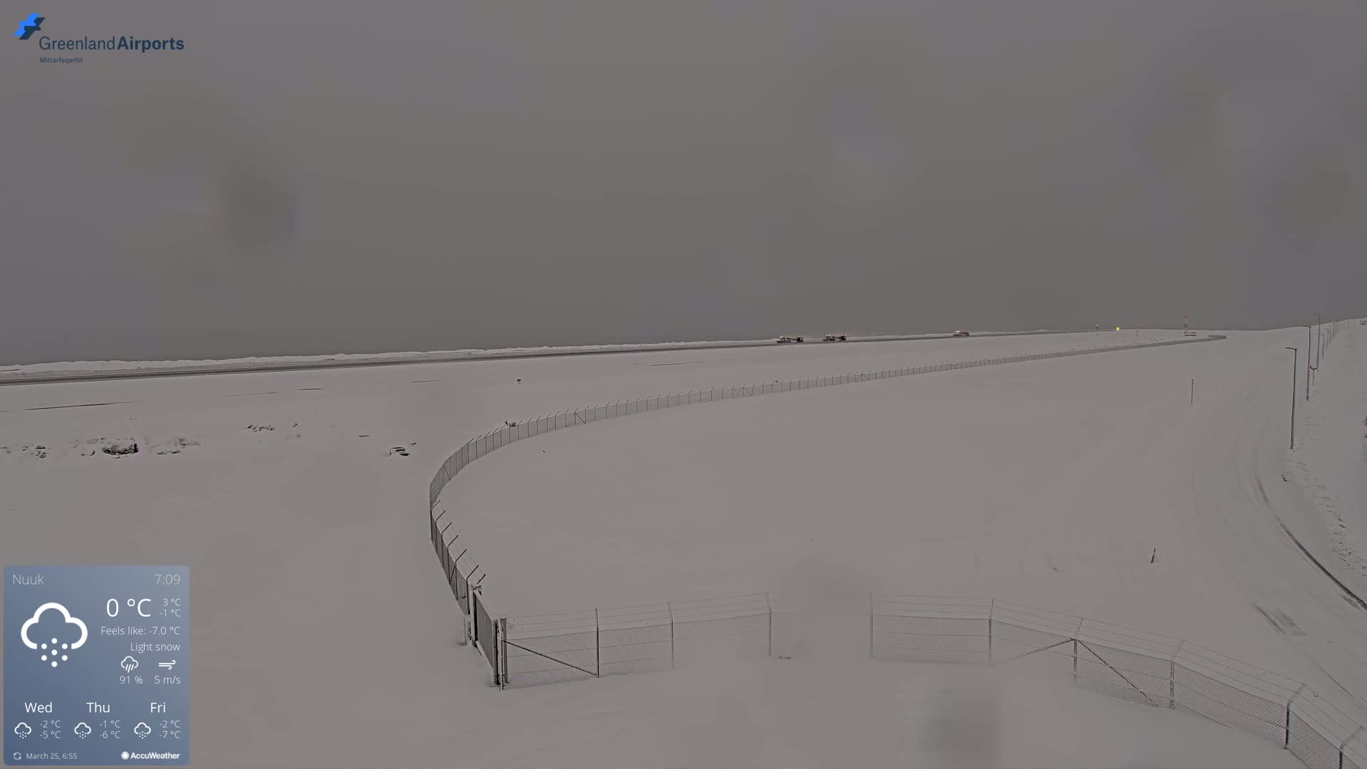 A snow-covered airport runway and surrounding area are visible under a gray, overcast sky with light snow falling.