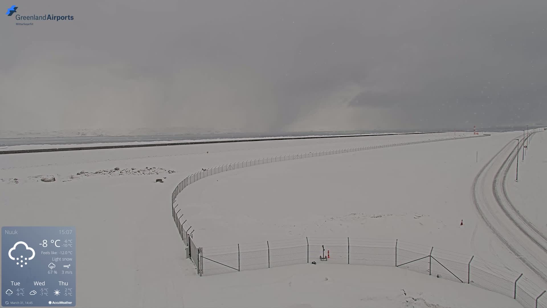 A snow-covered airfield with a chain-link fence under a gray, lightly snowing sky.