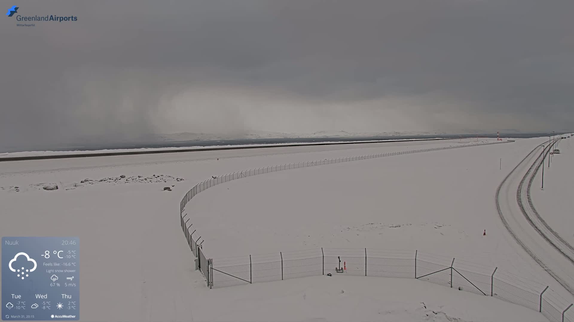 A snow-covered airfield with a chain-link fence and tire tracks in a light snow shower under an overcast sky.