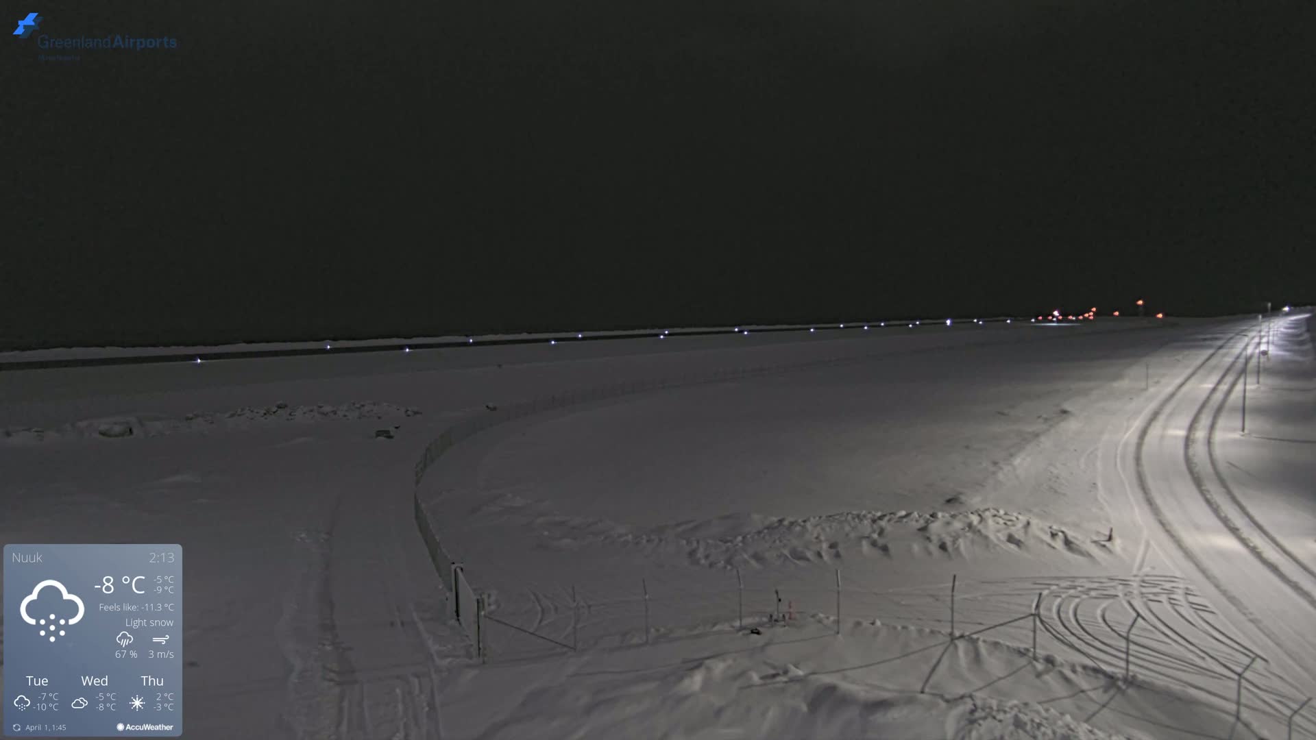 A snow-covered, dark landscape at night is illuminated by lights along a road and fence, with light snow falling.