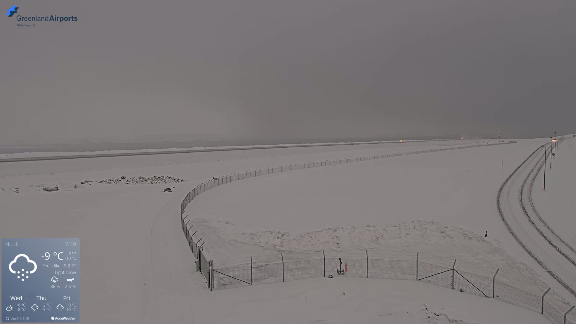 A snow-covered airport runway and surrounding area are visible under a light snow and overcast sky.