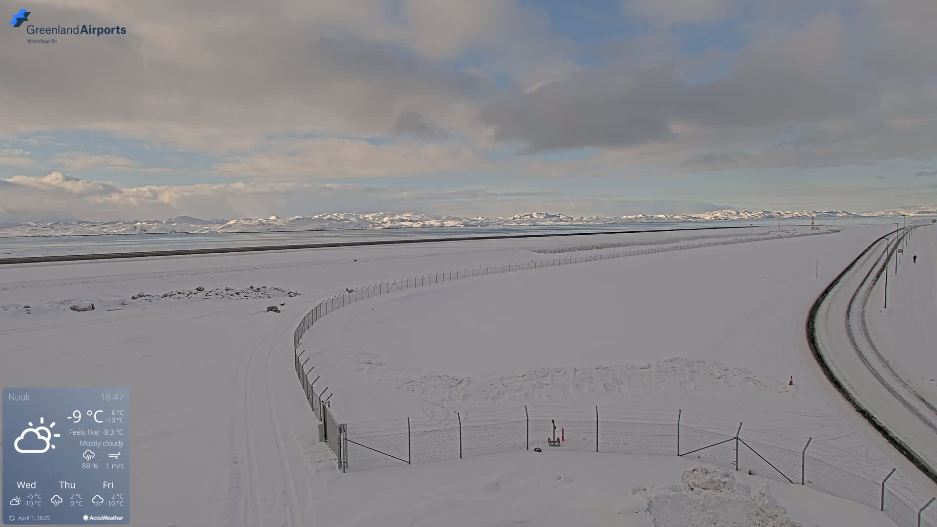 A snow-covered airport tarmac and runway extend towards a distant mountain range under a mostly cloudy sky.