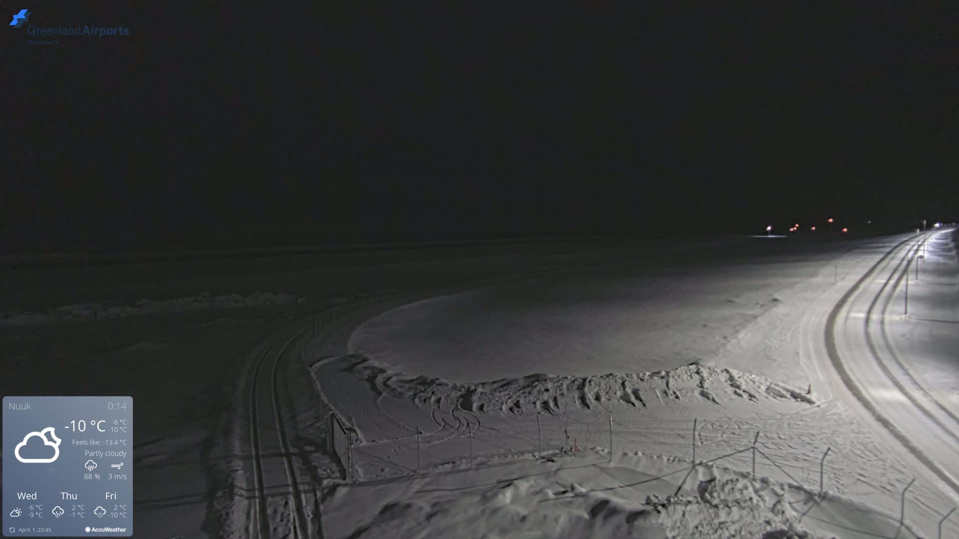 A snow-covered, nighttime landscape shows a road with tire tracks, lit by streetlights, next to a fenced-in, snow-covered area under a partly cloudy sky.