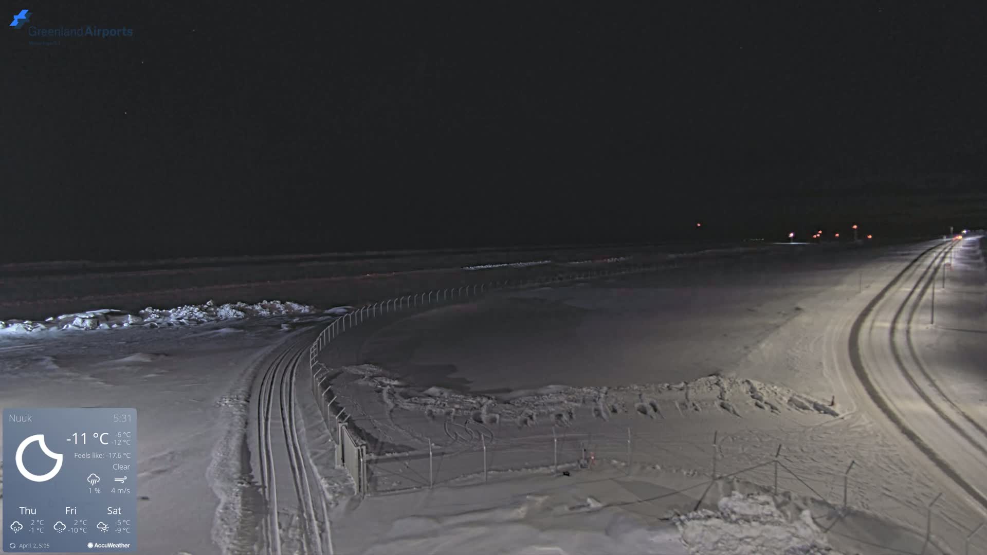 A snow-covered, nighttime scene shows a roadway curving away from the viewer, alongside a snow-covered field enclosed by a fence, under a clear sky.