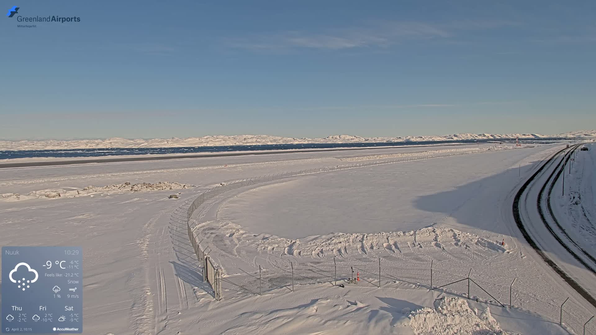 A snow-covered airport runway and road extend toward a frozen body of water and distant snow-capped mountains under a clear blue sky.