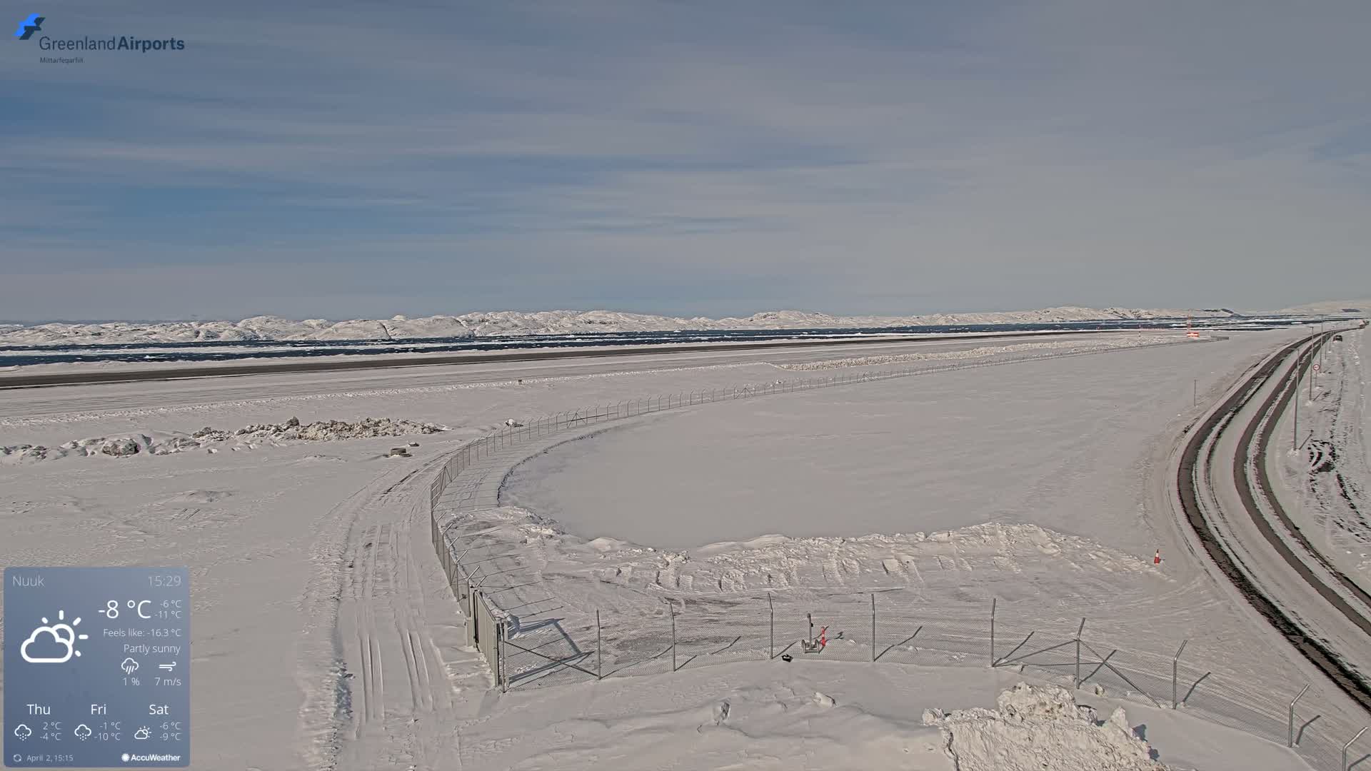 A snow-covered airport runway and surrounding area with partly sunny skies and snow-covered mountains in the distance.