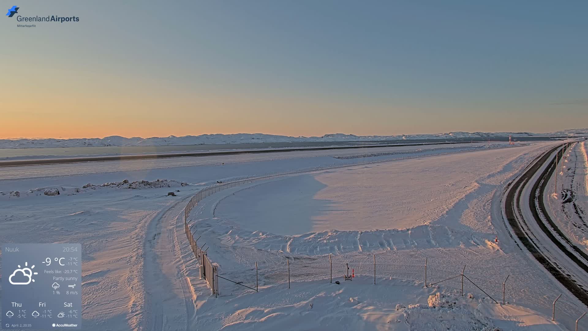 A snow-covered airport runway and road are visible under a partly sunny sky during sunset, with mountains in the background.