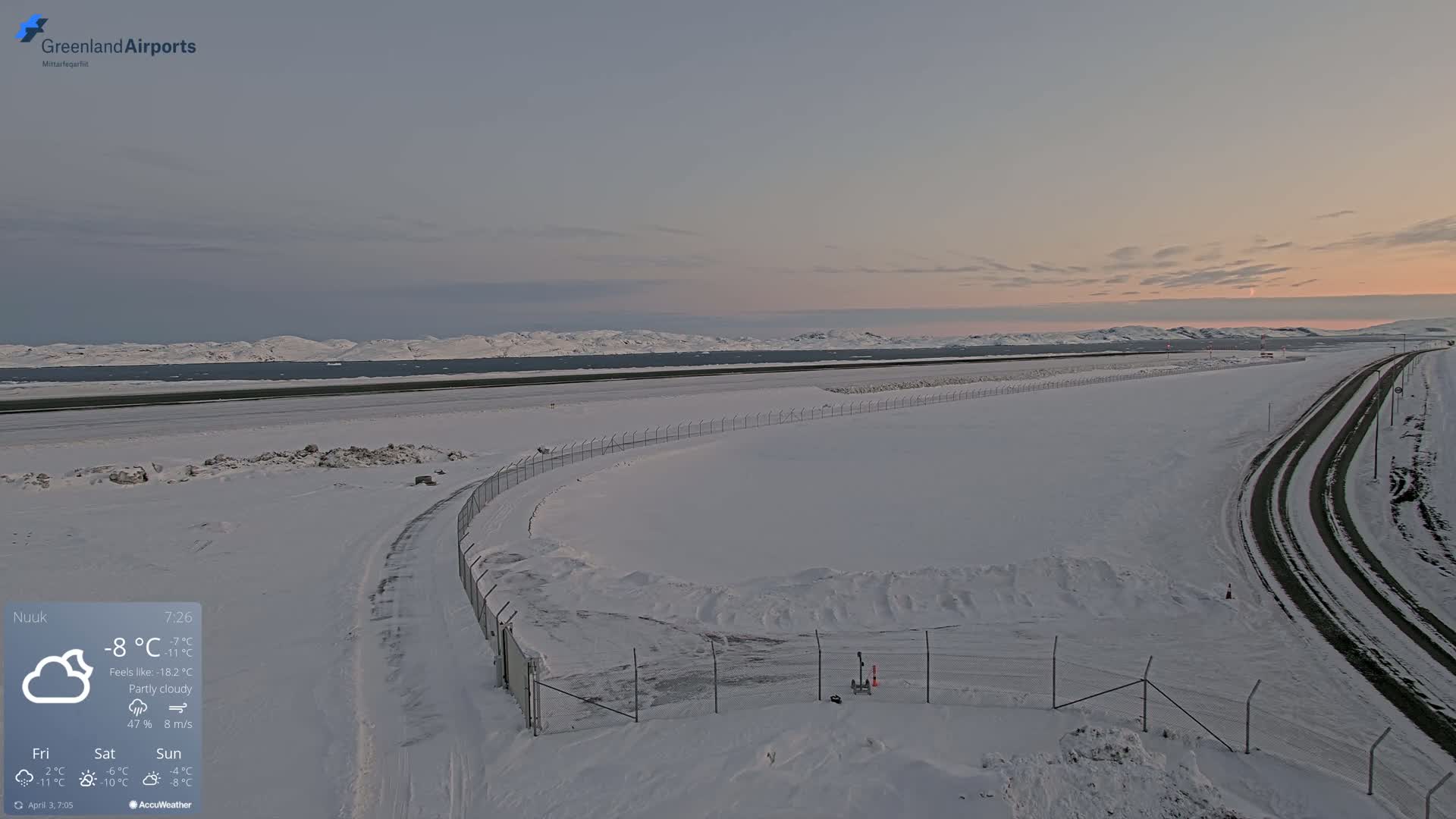 A snow-covered airport runway and access roads are visible under a partly cloudy sky at sunset, with mountains in the background.