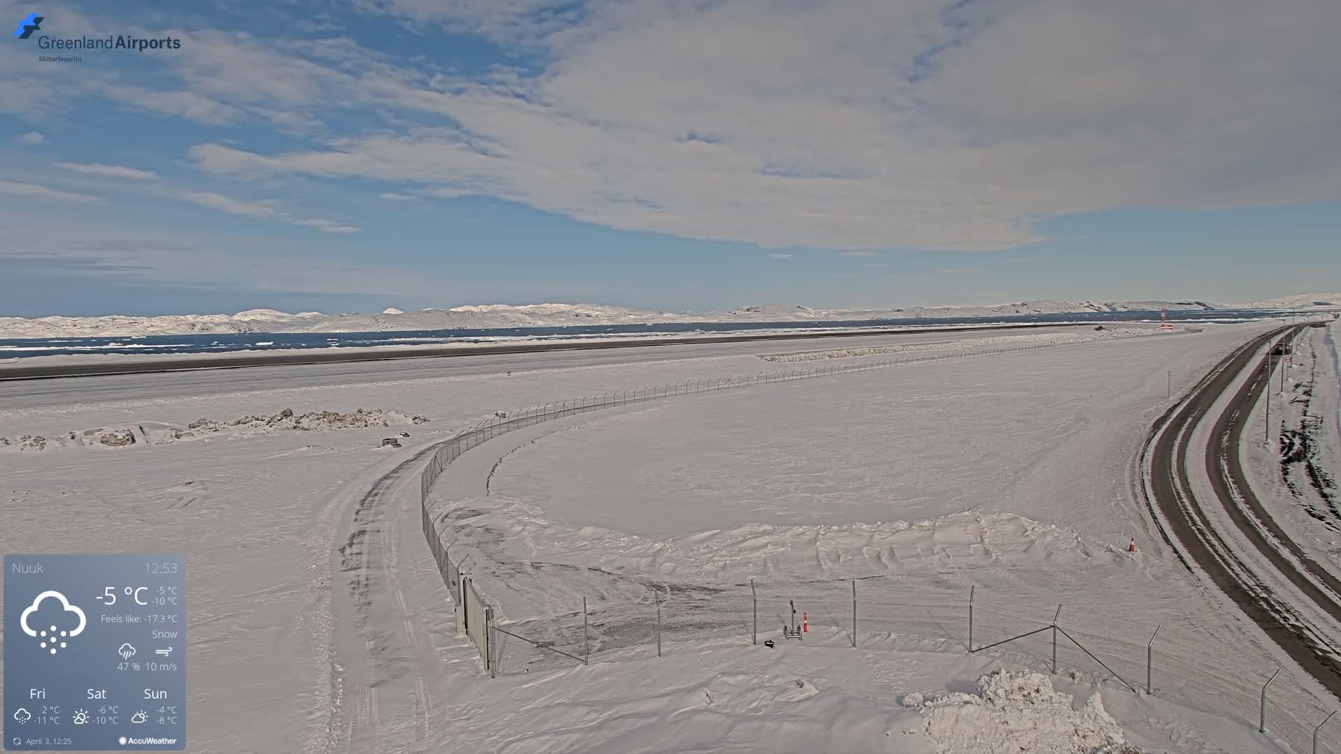 A snow-covered airport runway and surrounding area, with a partly cloudy sky and distant, snow-capped mountains visible in the background.