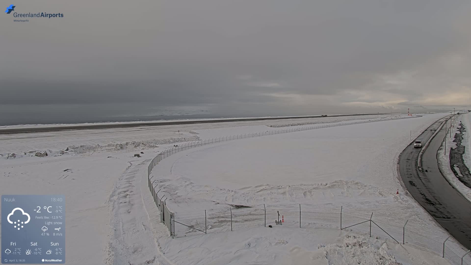 A snow-covered airport runway and access road are visible under a gray, overcast sky with light snow falling.