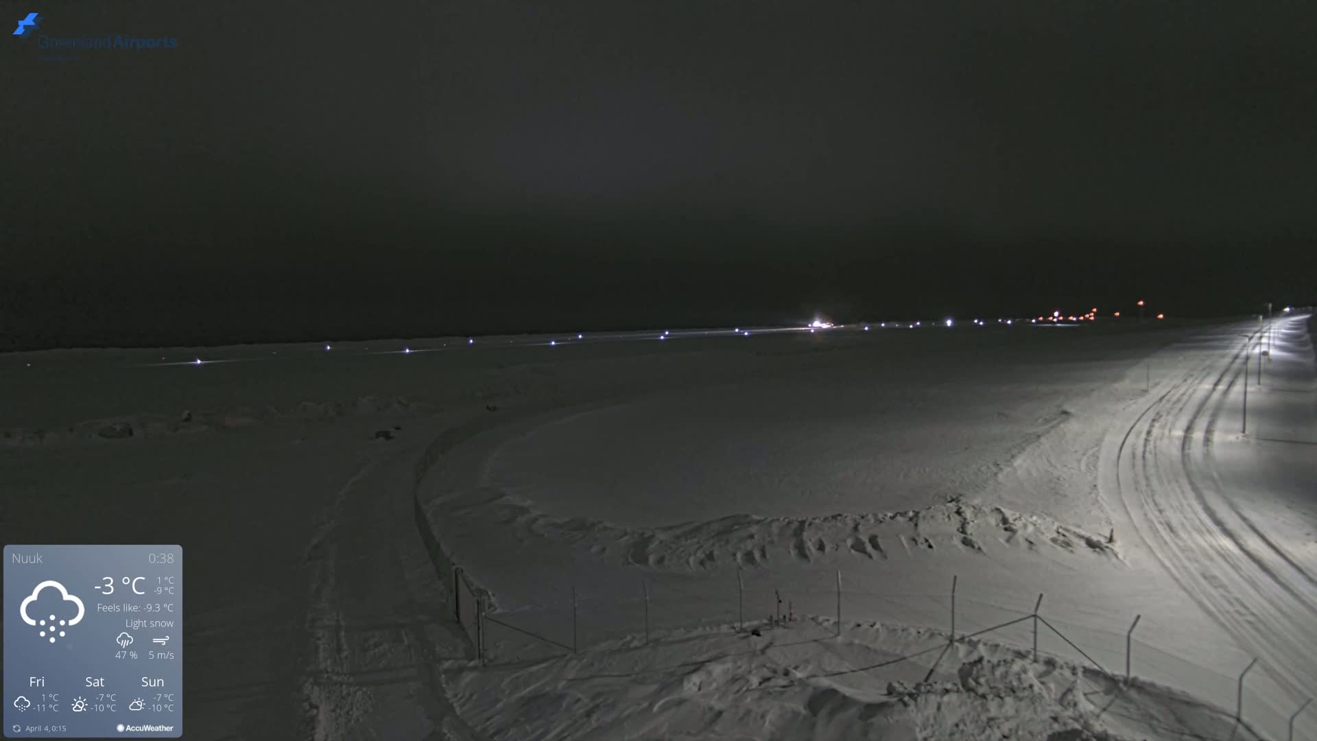 A snow-covered airfield at night during a light snow with faint lights illuminating the runway and surrounding area.
