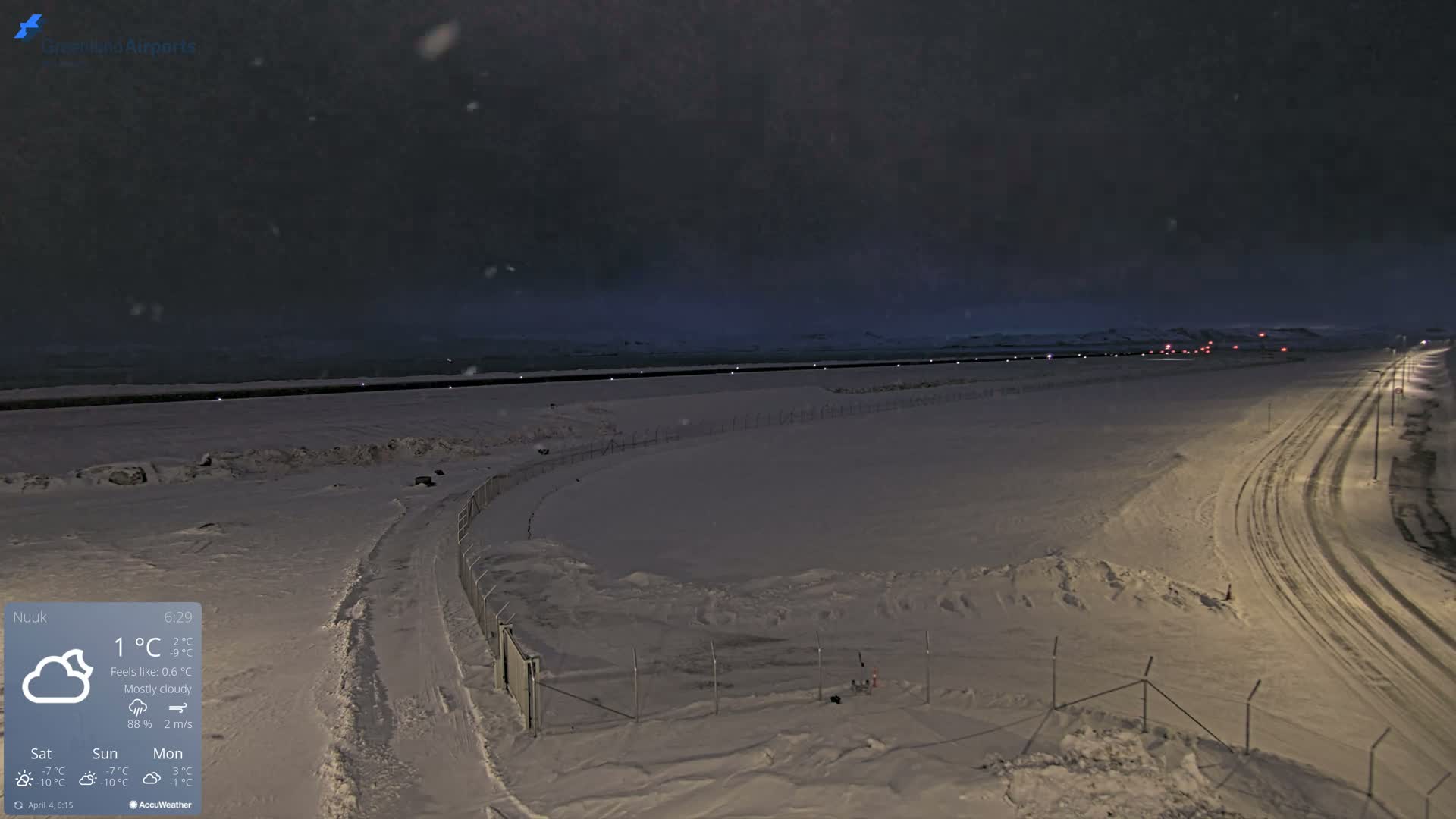 A snow-covered airport runway and surrounding area are visible under a mostly cloudy, nighttime sky with light snowfall.