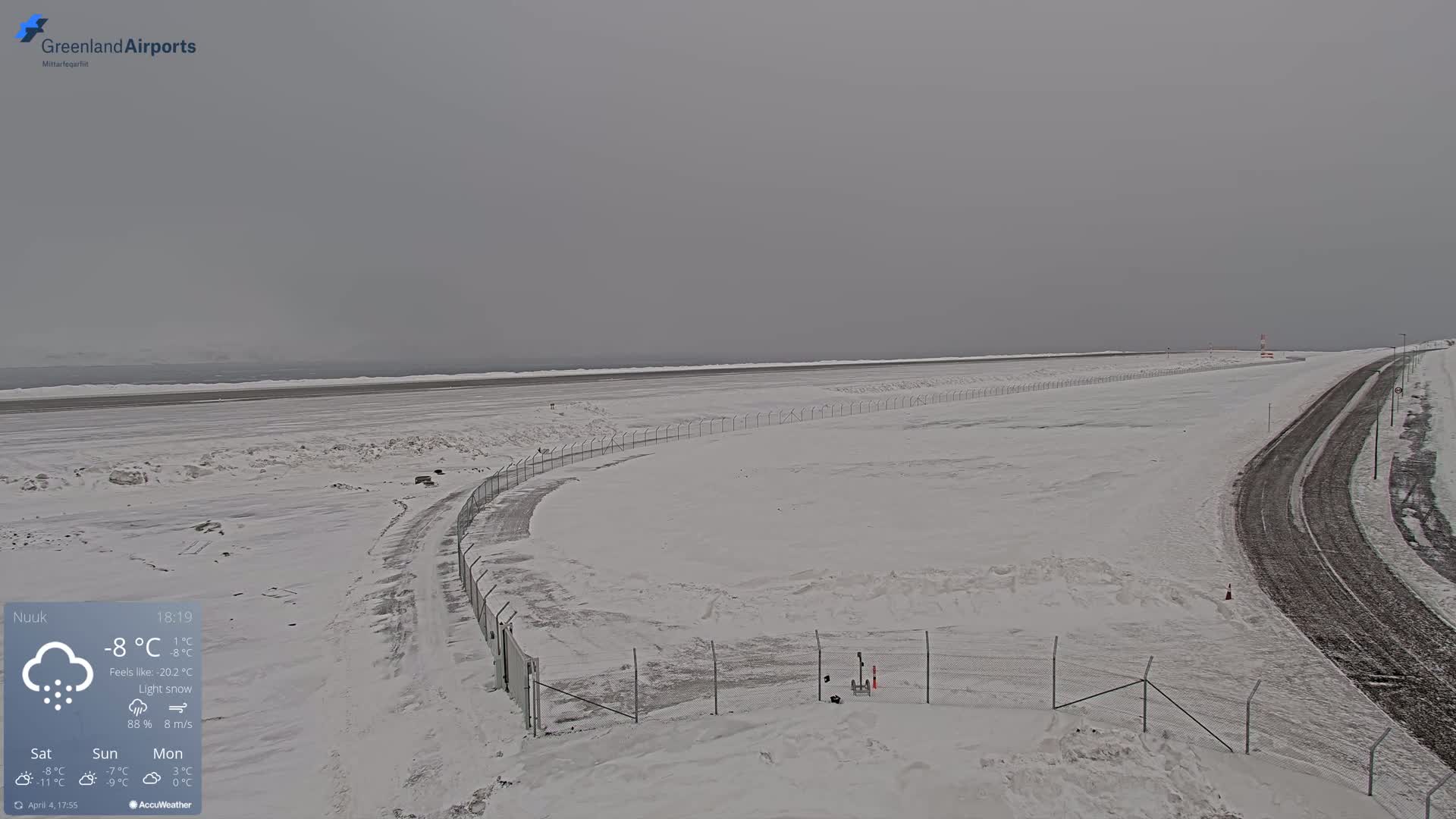A snow-covered airfield with a road beside it under a gray, overcast sky with light snow falling.