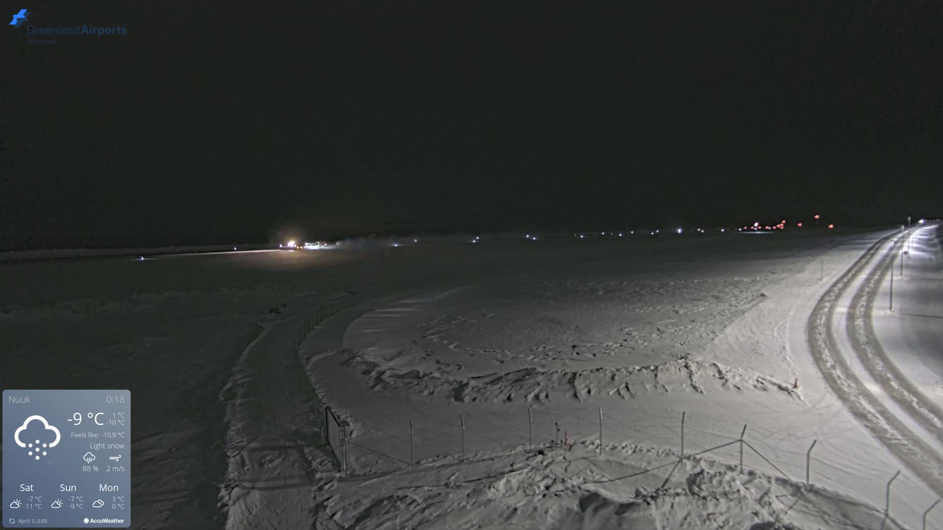 A snow-covered airfield at night under light snow with a few vehicles visible and a road with tire tracks leading away from the scene.