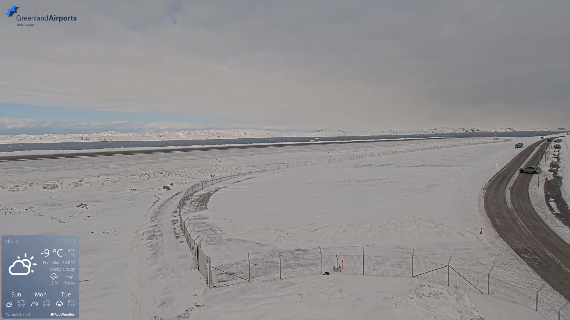 A snow-covered airport runway and road extend toward a mostly cloudy sky and distant, snow-capped mountains.