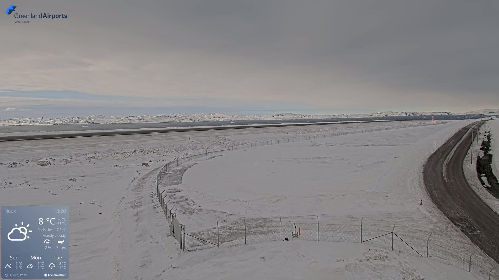 A mostly cloudy, snowy airfield with a road curving alongside a chain-link fence, overlooking a snow-covered body of water and distant mountains.