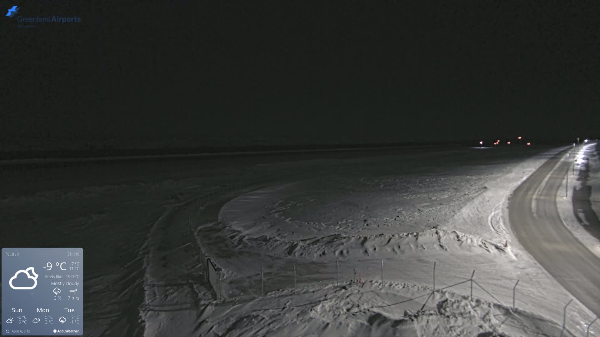 A snow-covered airfield and road are visible at night under mostly cloudy skies.