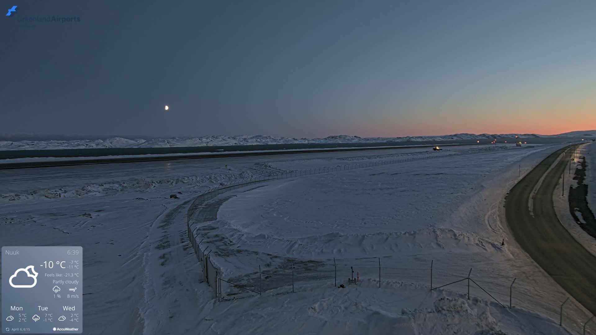 A snow-covered airport runway and adjacent road are visible at twilight under partly cloudy skies.