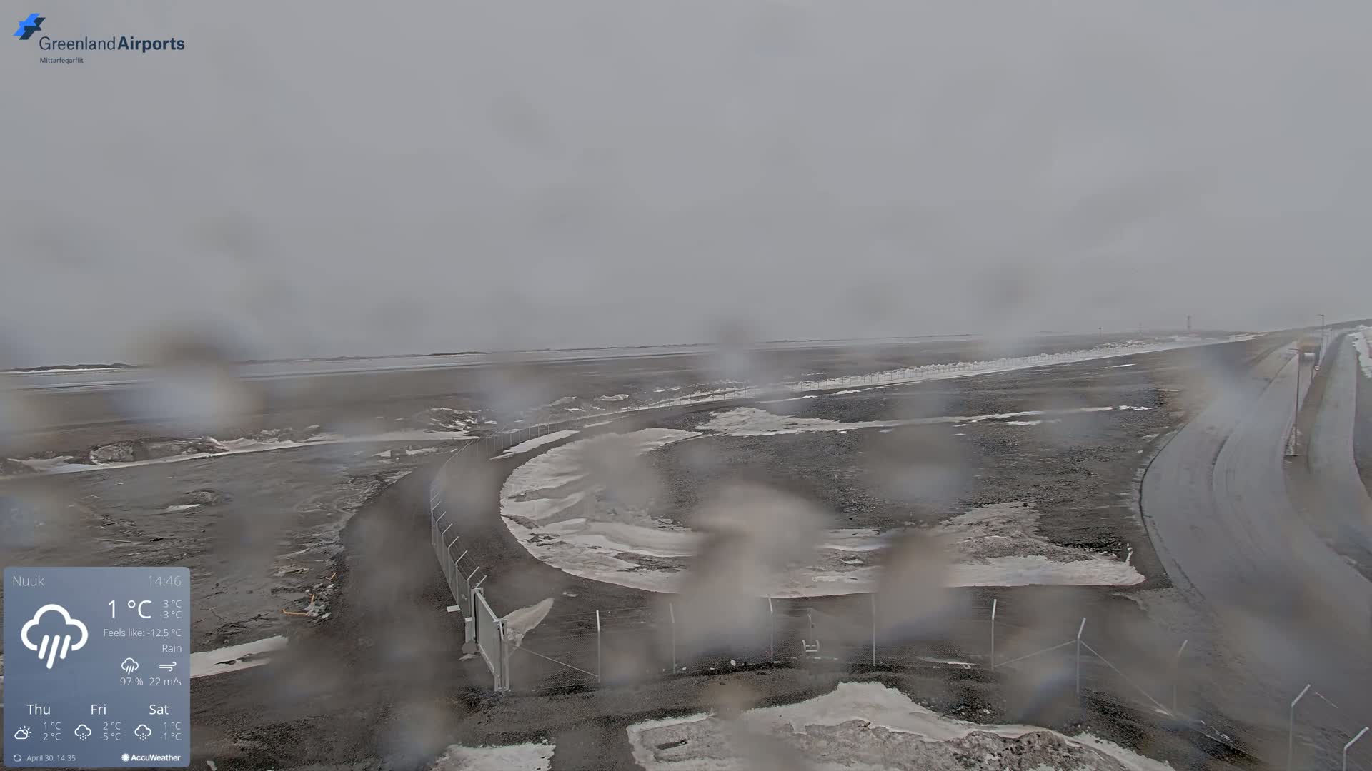 A rain-covered, overcast view of an airport tarmac with patches of snow and ice, and a fence surrounding a curved roadway.